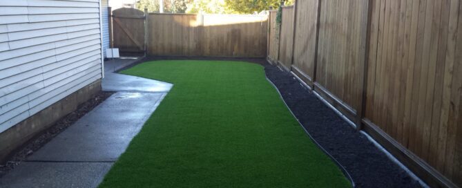 Installed artificial turf lawn in a narrow backyard space next to a concrete path and wood fence.