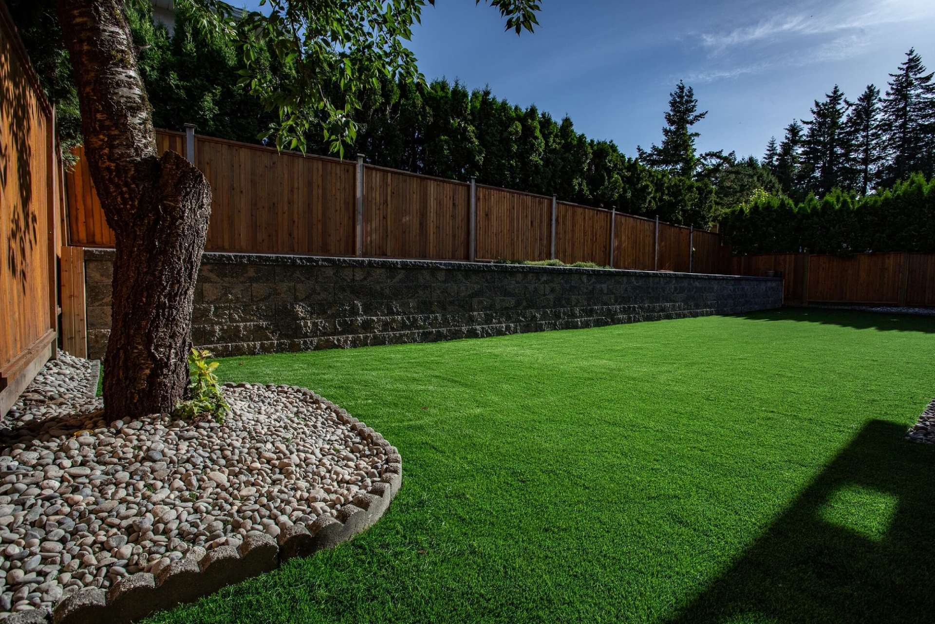 Artificial turf yard with stone retaining wall, privacy fence, river rock border, and large tree.